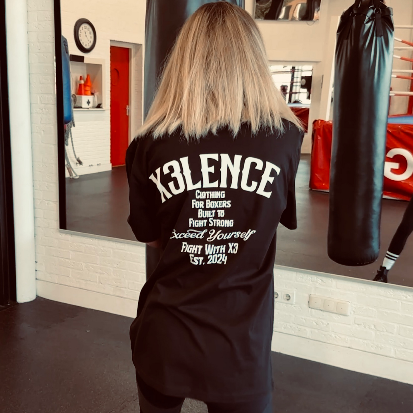 Vrouw draagt zwart X3LENCE Fight With X3 T-shirt met grote rugprint “Clothing for Boxers – Exceed Yourself”, gefotografeerd in een boksschool
